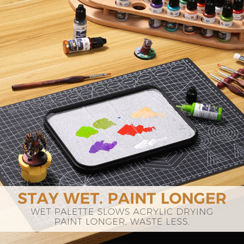 Wet palette with mixed acrylic paints on hydro paper alongside hobby tools, paint bottles, and painted miniature.